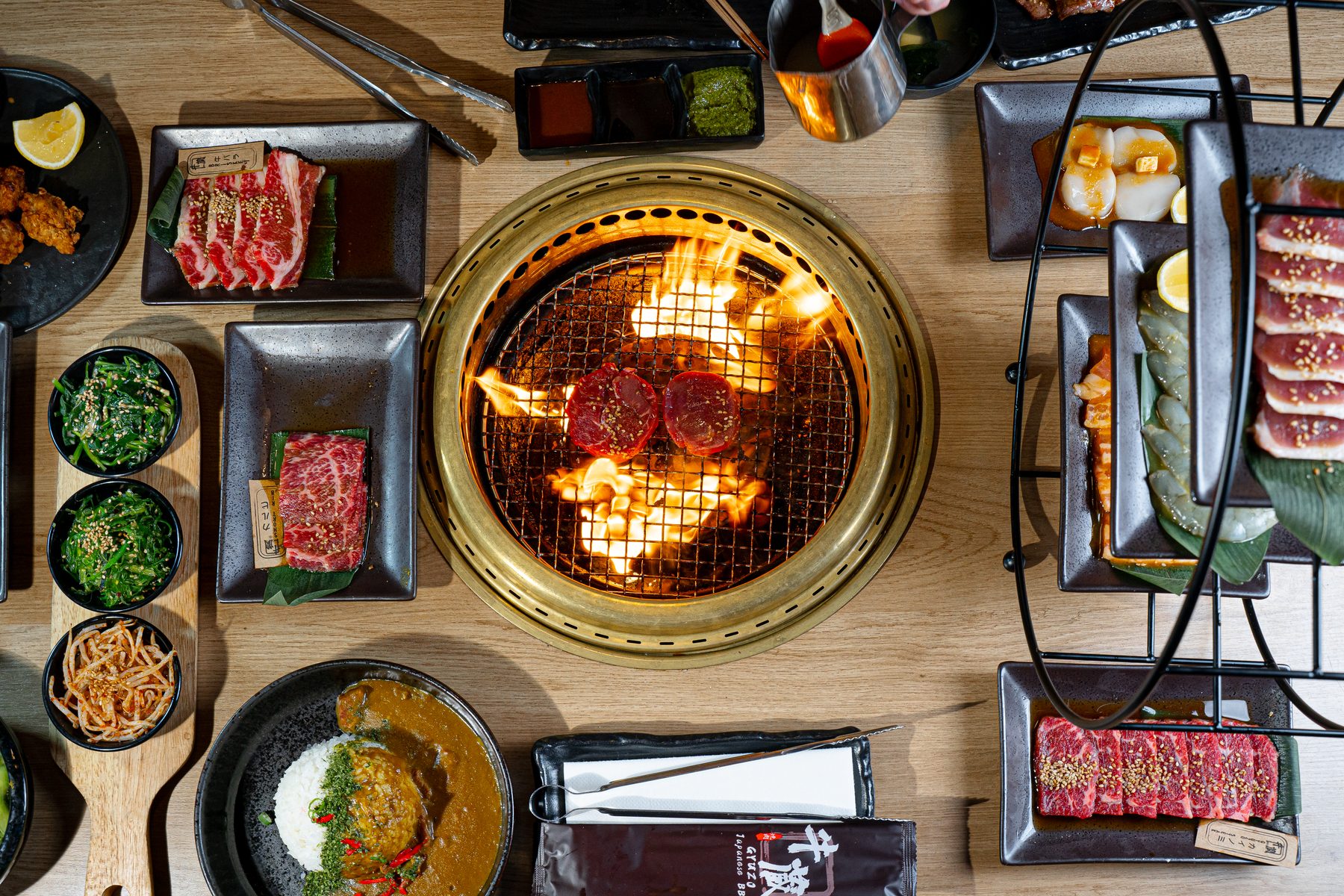 Overhead view of a brass tabletop grill with flames under two slices of beef, surrounded by plates of marbled Wagyu, sliced meats and seafood at Gyuzo BBQ