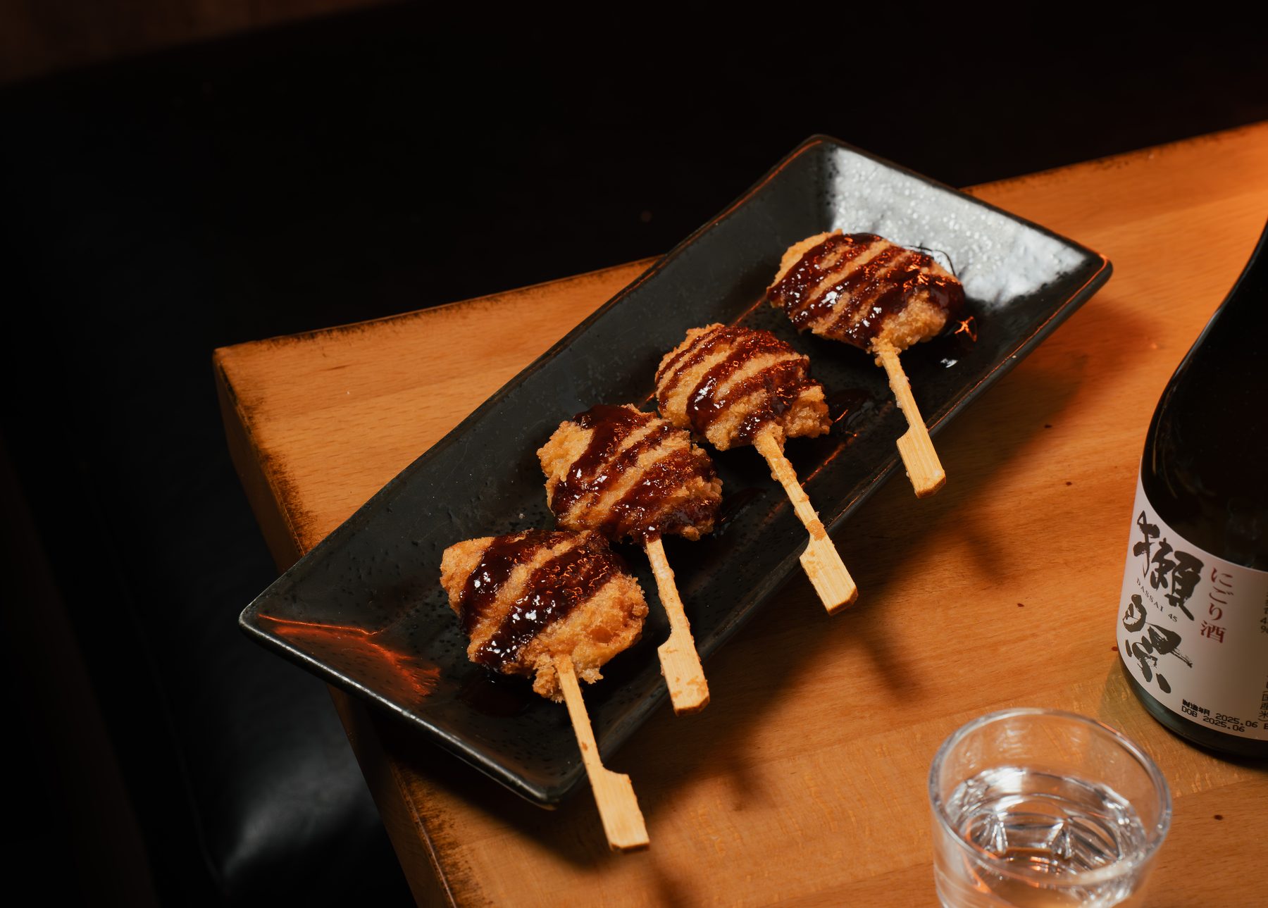 Four breaded, deep-fried kushiage skewers drizzled with dark sauce on a black plate beside a bottle of Dassai sake at Izakaya 68