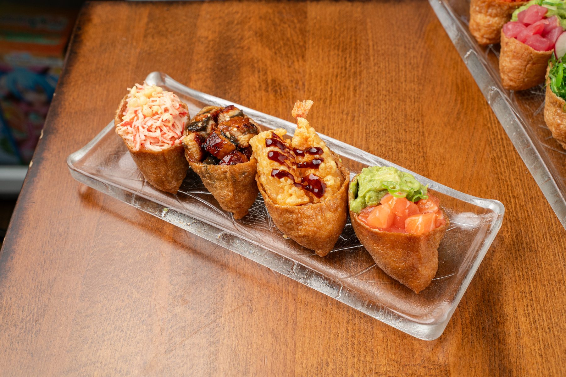 Four inari sushi topped with crab salad, eel, shrimp tempura and salmon with avocado on a glass tray at Izakaya Nana