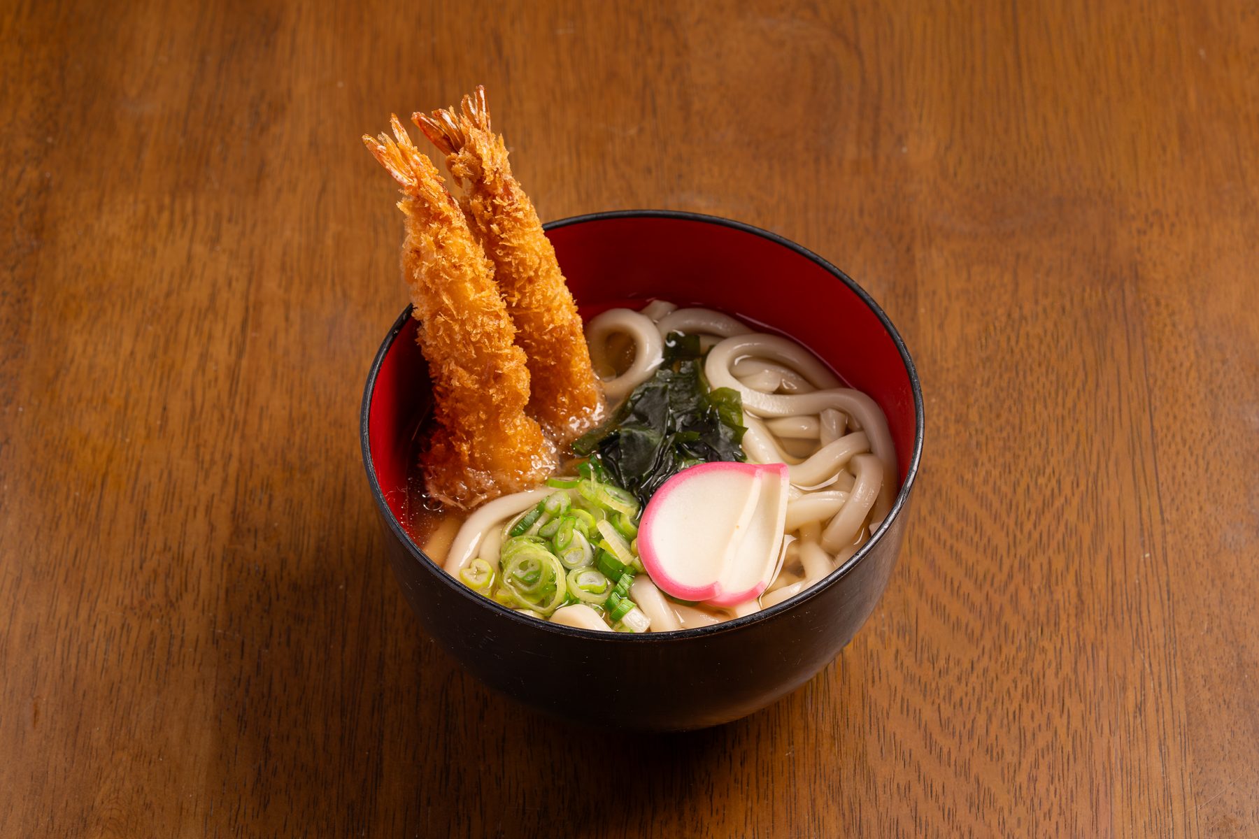 Shrimp tempura udon with two fried shrimp, wakame, a kamaboko slice and scallions in a black lacquer bowl at Okaeri Japanese Cafe