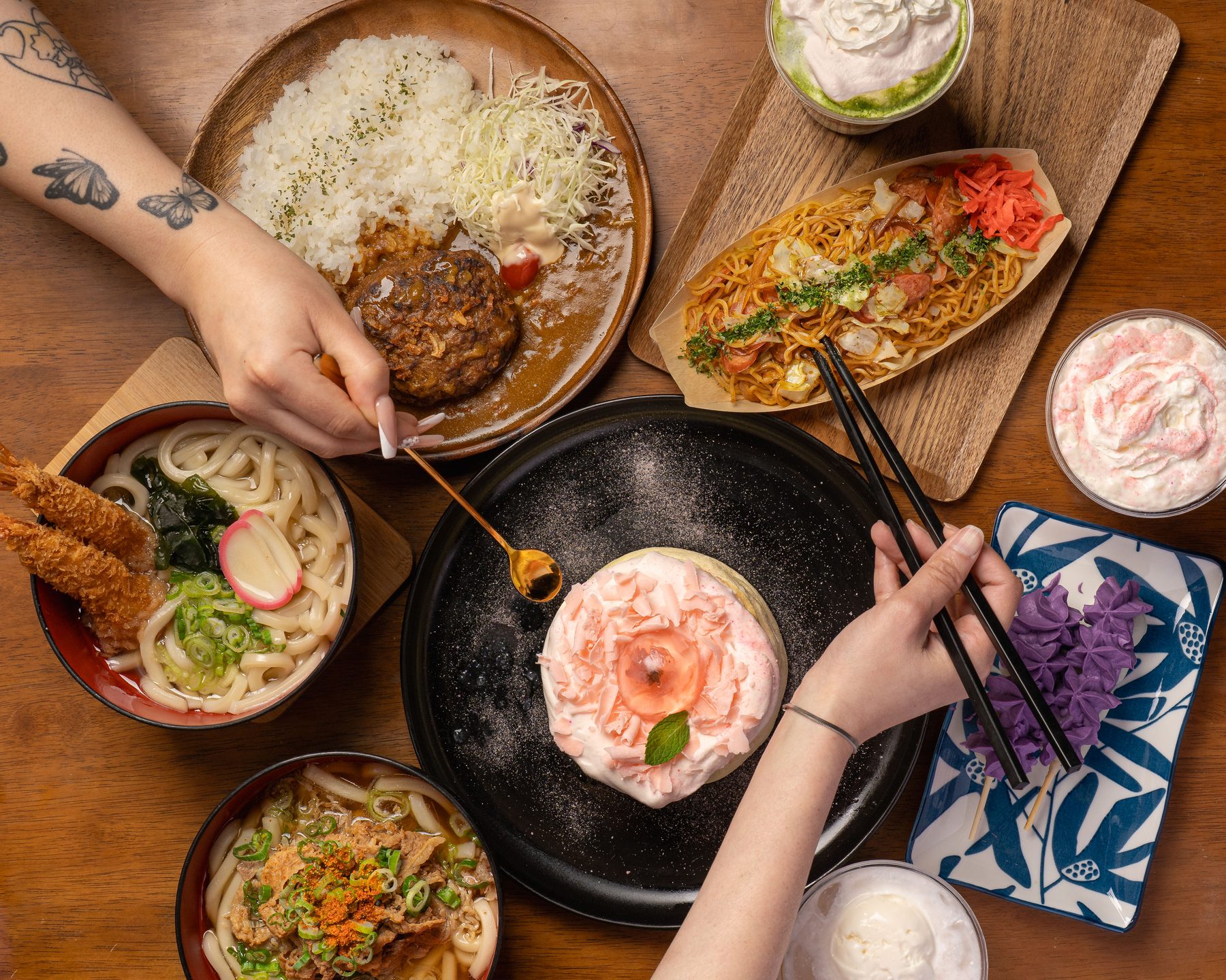 Overhead table of udon, curry with hamburg steak, yakisoba, ube dango and a pink soufflé pancake in the center at Okaeri